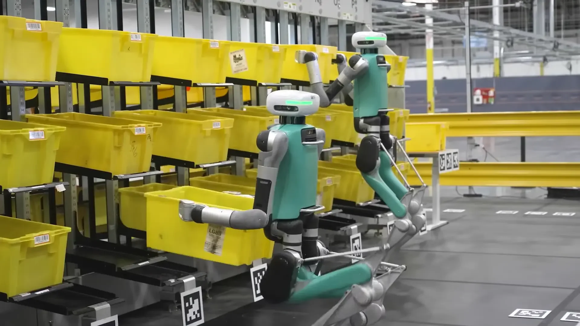 Humanoid robots working in a logistics warehouse, autonomously moving and sorting yellow bins on shelves.