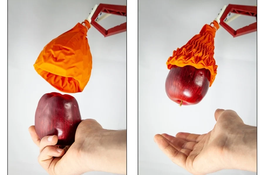 Soft robotic gripper shaped like a balloon gently wrapping around and lifting a red apple, showing ability to handle delicate objects.