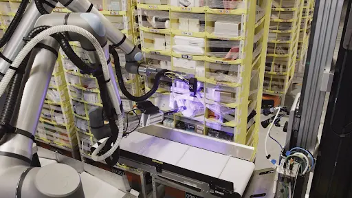 An automated robotic arm in a warehouse uses computer vision to pick items from shelves for order fulfillment on a conveyor belt