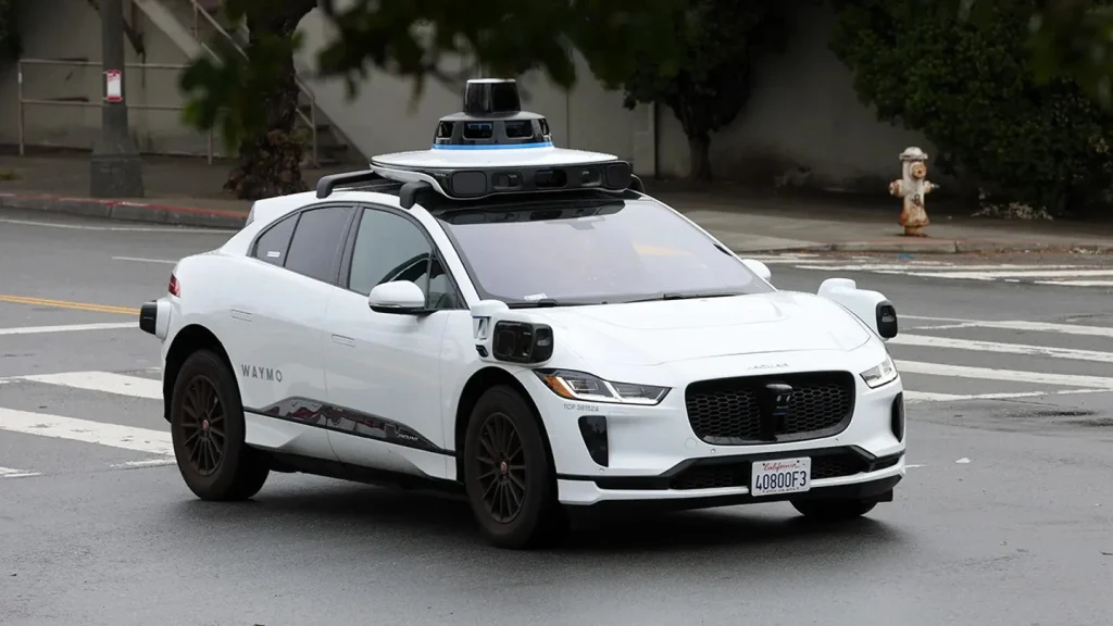 A white Waymo self-driving car, a Jaguar I-PACE, navigates a city street using its rooftop Lidar and camera sensors