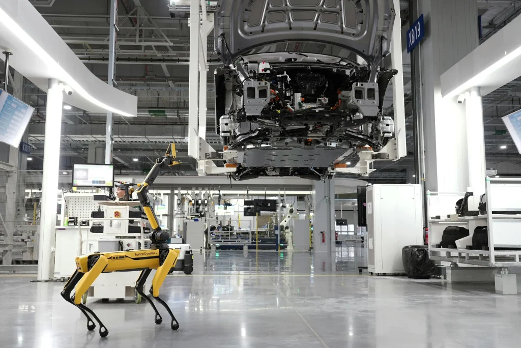 The Boston Dynamics Spot robot with an arm attachment inspecting a modern automotive factory, walking under a car chassis