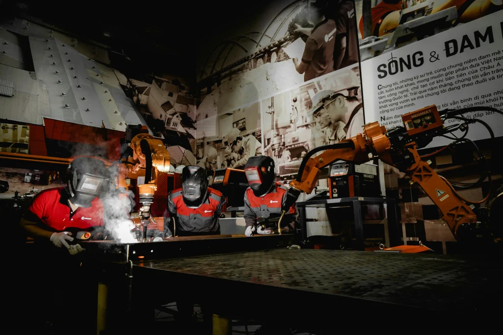 Welders in protective gear work alongside a large orange robotic arm in a training facility, demonstrating human-robot collaboration