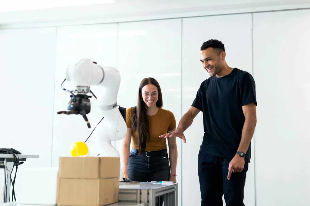 Two people smiling as they interact with a white collaborative robot (cobot) in a lab, showcasing safe human-robot interaction