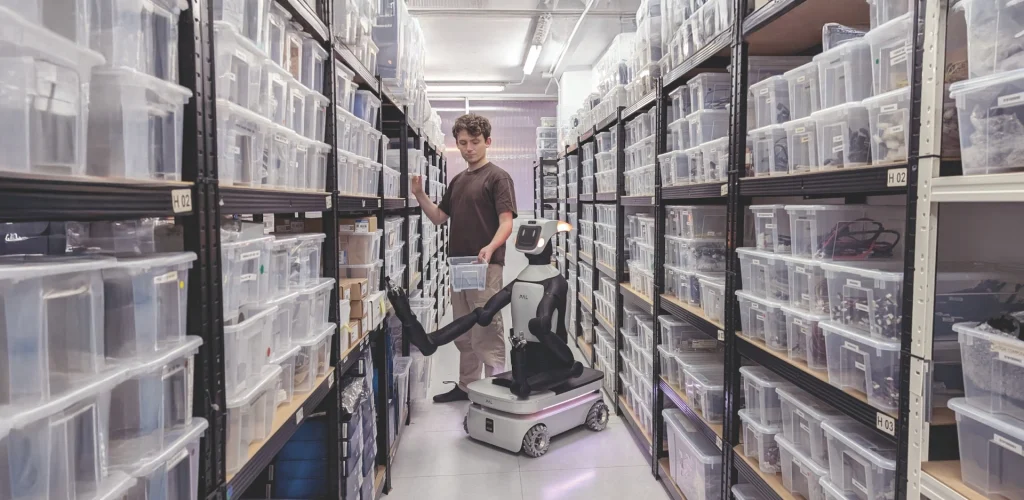 A man and a collaborative mobile robot work together in a narrow warehouse aisle, with the robot handing him a plastic bin