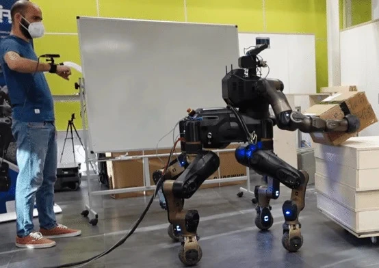 A four-legged robot with two arms, teleoperated by a person, carries a cardboard box in a warehouse demonstration