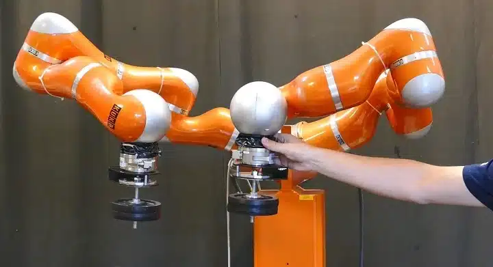 A human hand guides a triple-armed orange collaborative robot during a kinesthetic training demonstration