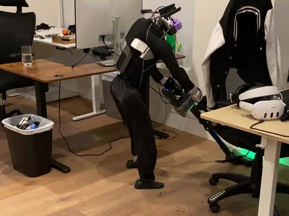 A humanoid robot holding a handheld vacuum cleaner to clean the floor under an office desk