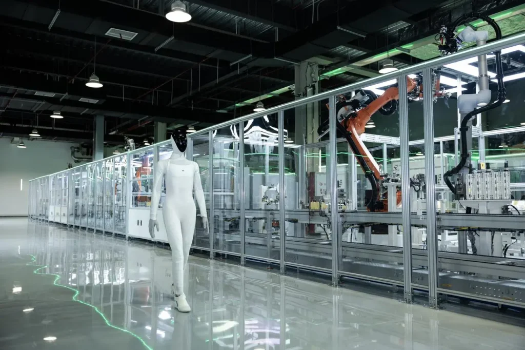 A sleek white humanoid robot walking past automated robotic cells in a modern production facility