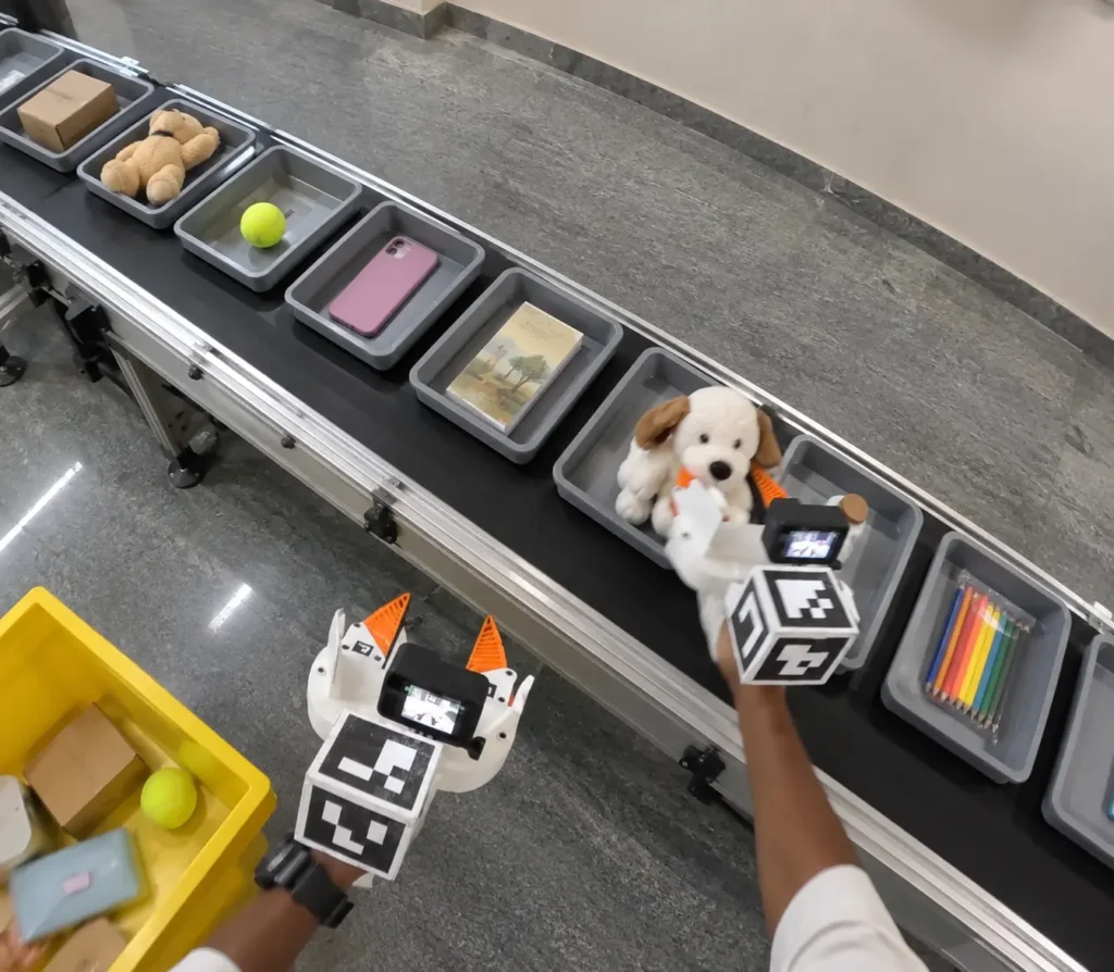 First-person view of an operator using tracked gripper rigs to collect object-sorting data on a conveyor belt